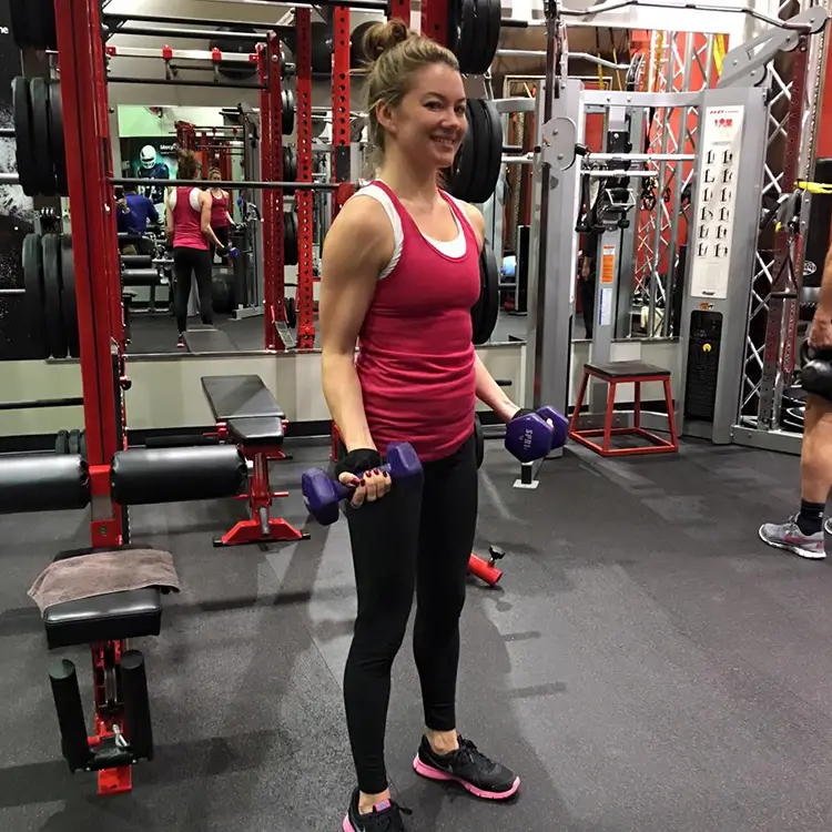 A woman in a pink tank top and black leggings is standing in a gym holding purple dumbbells. She is smiling and appears to be exercising. Gym equipment is visible in the background.