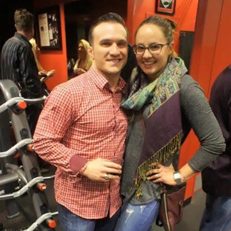 A man and a woman pose together indoors near a red wall. The man wears a red checkered shirt, and the woman wears glasses, a scarf, and a gray top. Exercise equipment is visible in the background.