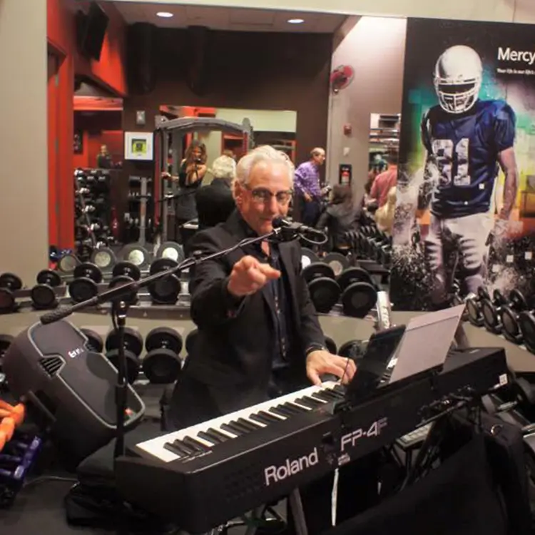 Man playing Roland FP-4 keyboard and singing into a microphone in a gym with exercise equipment and a football poster in the background.