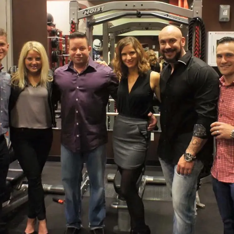 Group of six people standing and posing for a photo in a gym. They are dressed in casual and semi-formal attire. Workout equipment is visible in the background.