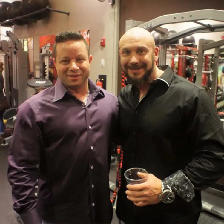 Two men are posing for a photo at a gym. Both are wearing dark long-sleeve shirts; one man holds a drink. Exercise equipment is visible in the background.