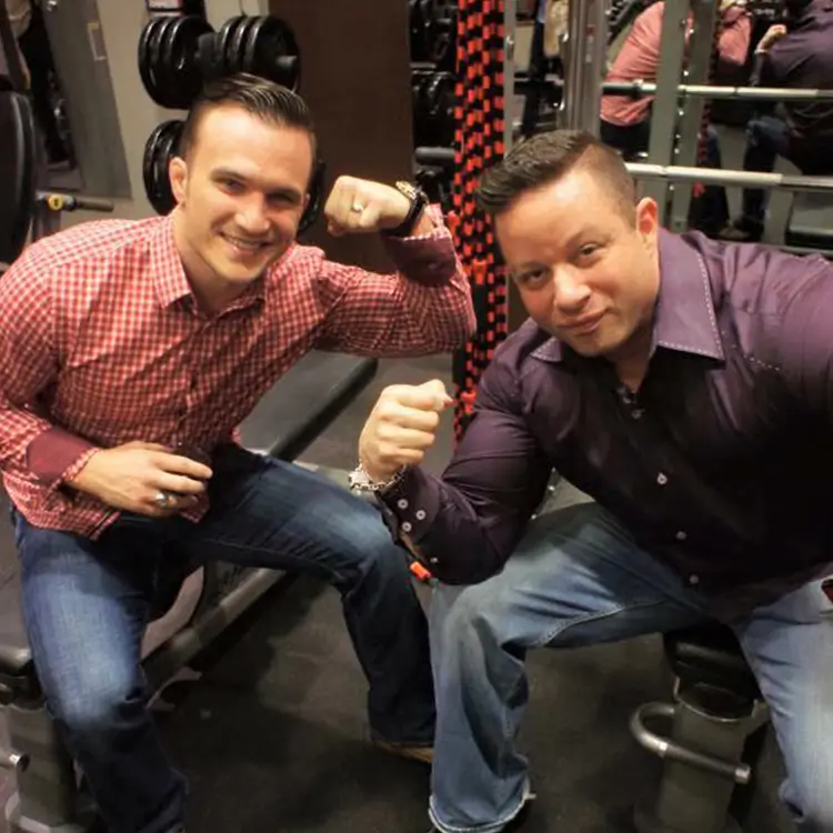 Two men seated in a gym, both flexing their biceps and smiling at the camera. The man on the left wears a red checkered shirt, and the man on the right wears a dark-colored shirt.