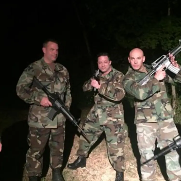 Three individuals in military camouflage uniforms pose with firearms in an outdoor setting at night, with Jamie Mushlin, the owner of Red Fitness Lounge, standing confidently in the center.