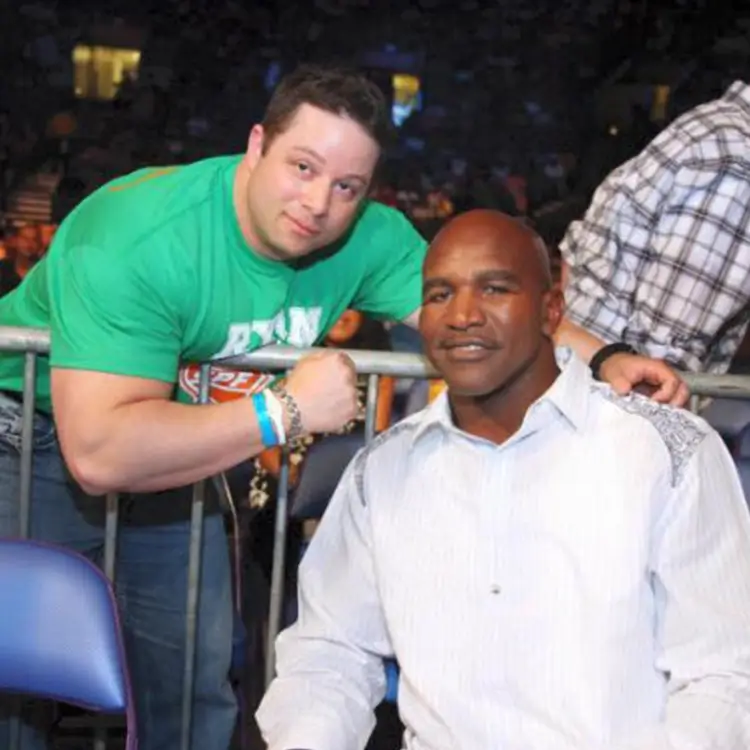 Two men pose for a photo together; one stands behind a barrier in a green shirt flexing his arm, while the other sits in front of the barrier wearing a white shirt.