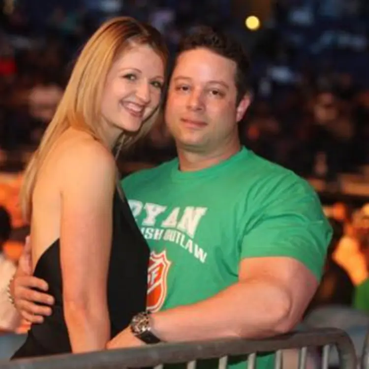 Jamie Mushlin stands with his girlfriend, both smiling, as he wraps his arm around her waist. They are in a public setting with people in the background, and Jamie wears a green T-shirt.