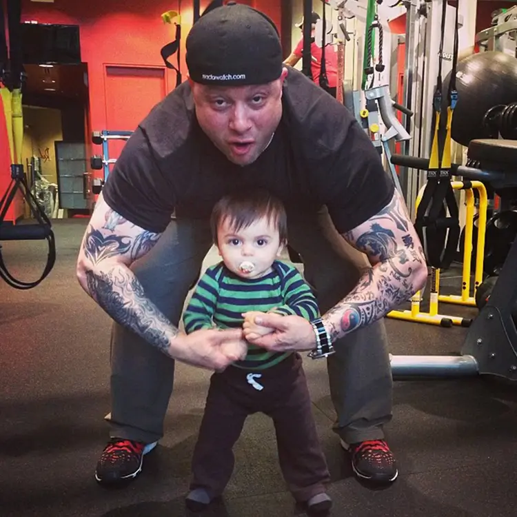 A man in a black cap and tattoos, who appears to be Jamie Mushlin the gym owner, squats behind a baby with a pacifier, holding the baby's hands, having fun together inside a gym with exercise equipment.