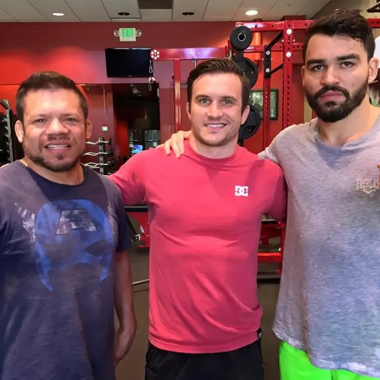 Three men standing in a gym, smiling at the camera. The man on the left is wearing a blue shirt, the man in the center is wearing a red shirt, and the man on the right is wearing a grey shirt and green shorts.