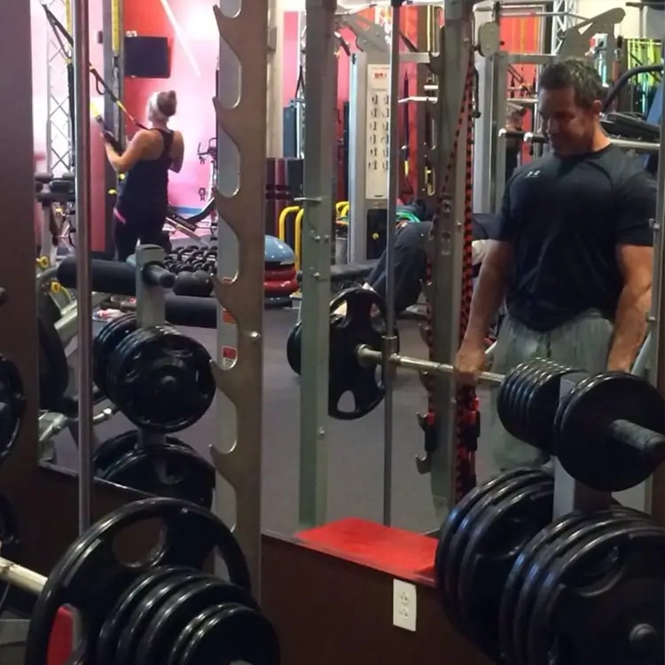 People are working out in a gym. A man with a barbell and a woman using a suspension trainer are visible, along with various gym equipment.