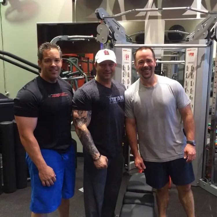 Three men standing in a gym, smiling at the camera. They are in front of exercise equipment, wearing workout attire including t-shirts, shorts, and a baseball cap.