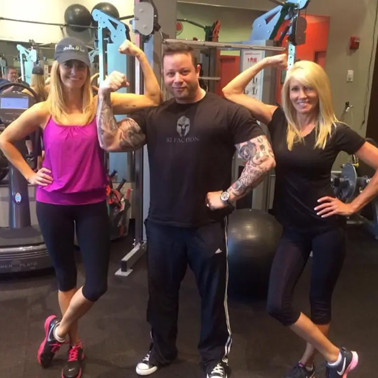 Two women posing with a man in a gym. The man is flexing his arms with tattoos visible, while the women stand on either side, smiling and mimicking a flexing pose. A gym setup is in the background.