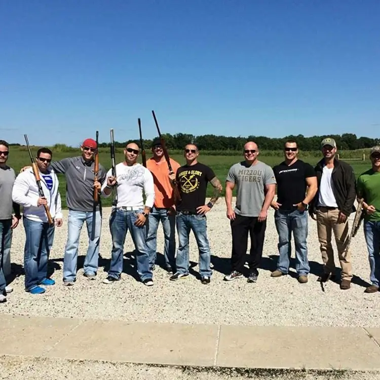 A group of men holding shotguns stands outdoors on a gravel area with clear skies and greenery in the background.