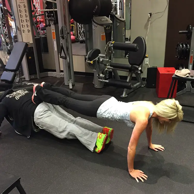 Two people are performing push-ups in a gym, where one person has their feet resting on the back of the other person. Equipment is visible in the background.