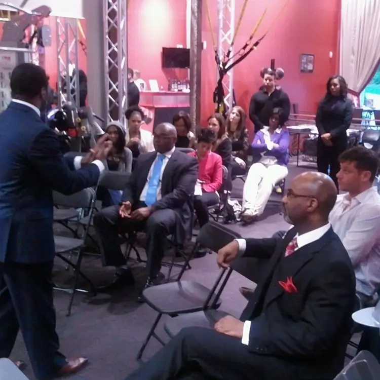 A person in a suit is giving a presentation to an audience seated on folding chairs in a room with red walls and large windows.