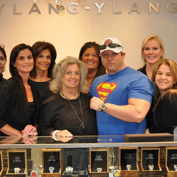 A group of people stand behind a glass display case with various watches on display. One person in the center is wearing a blue Superman shirt and a cap, posing with a woman in front of them.