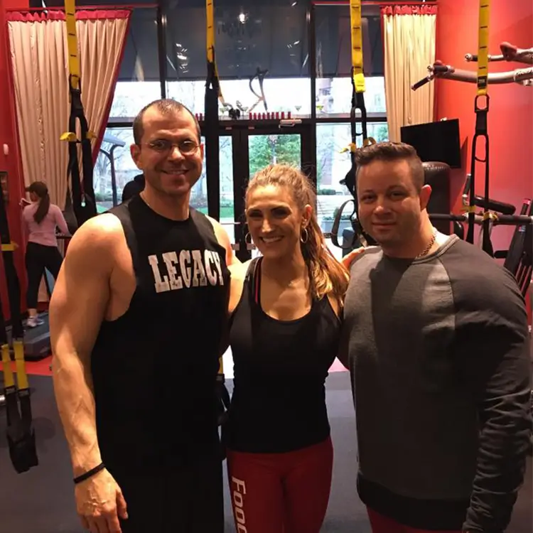 Three people posing together in a gym with exercise equipment and TRX suspension straps visible in the background.