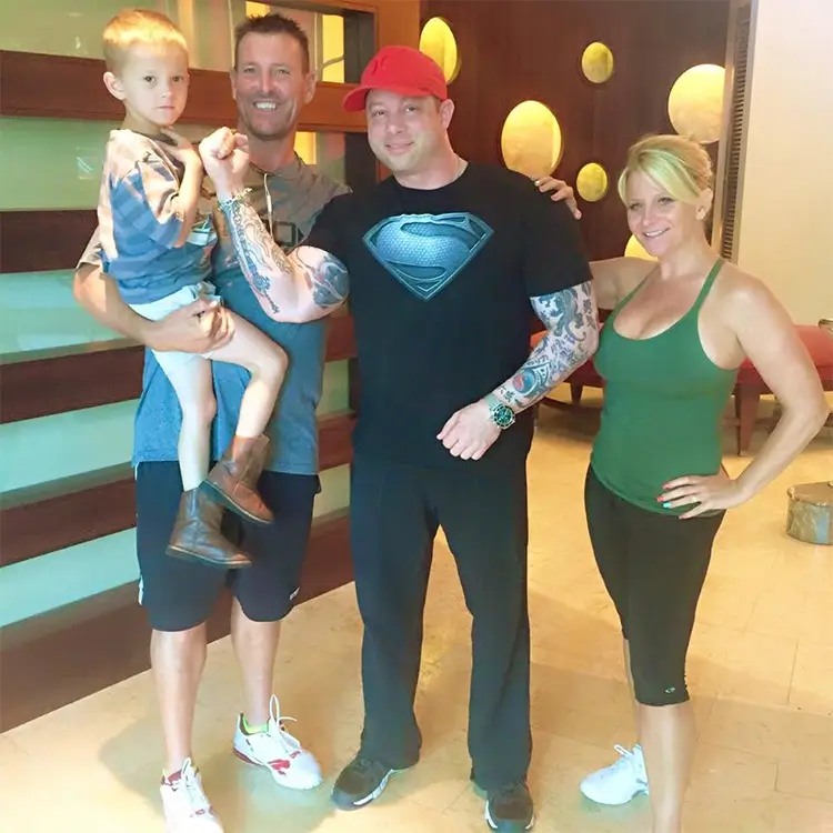 A group of four people, including a child, pose indoors. Three adults are in athletic attire, and one man holds the child. The man in the center wears a black Superman-themed shirt and a red cap.