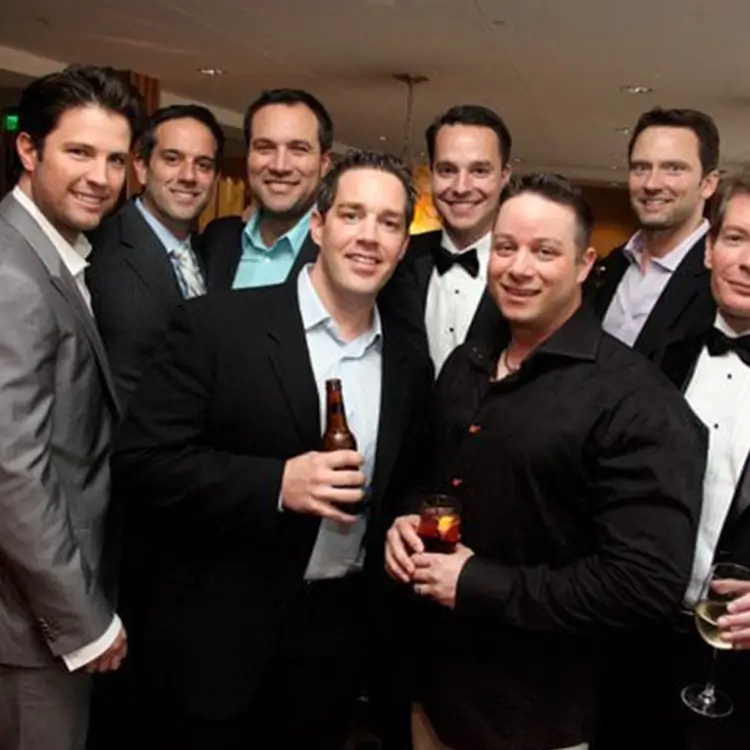 A group of men in formal and semi-formal attire pose together at an indoor event, with some holding drinks.