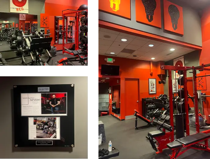 Compact gym with red and black decor. Equipment includes dumbbells, squat racks, and benches. A framed certificate and photo are mounted on the wall. Overhead lighting and artwork are visible.