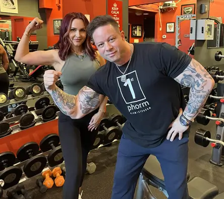 Two people posing and flexing their muscles in a gym with weights and equipment in the background.