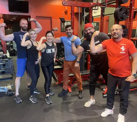 A group of six people in gym attire pose flexing their muscles in a gym setting with equipment in the background.