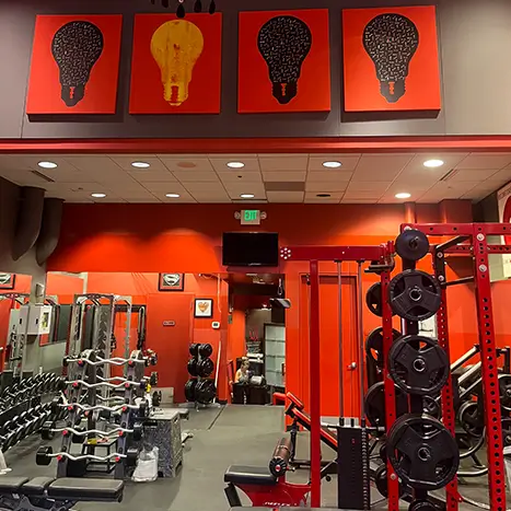 A gym interior with red and black decor, various weightlifting equipment, and three artwork pieces of light bulbs on the wall above.