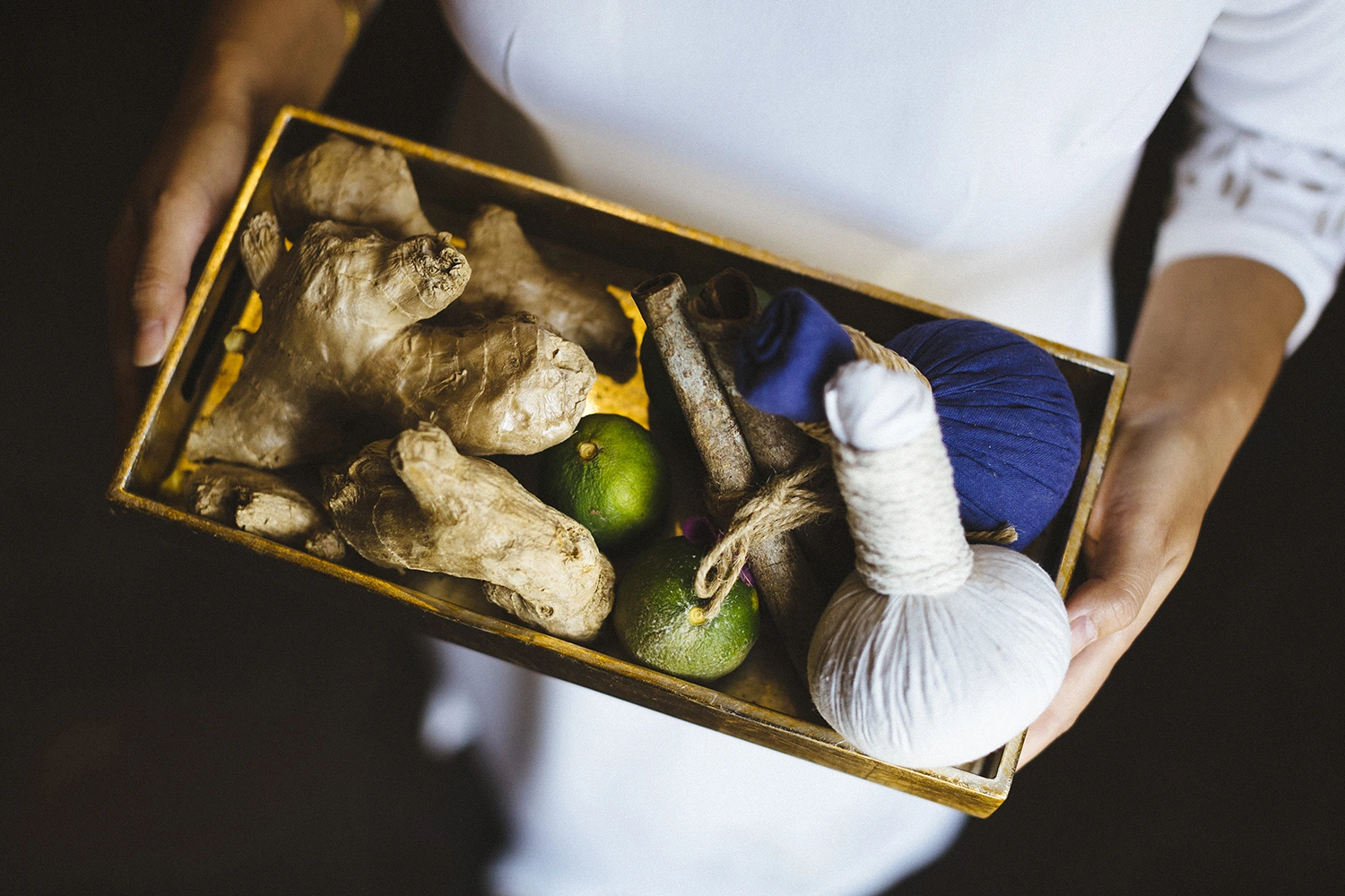 A person in white holding a wooden tray filled with spa items, including ginger roots, small green limes, cinnamon sticks, and white and blue herbal compress balls.