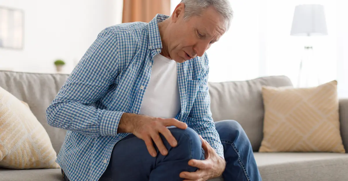 An older man sitting on a couch, holding his knee with a pained expression.