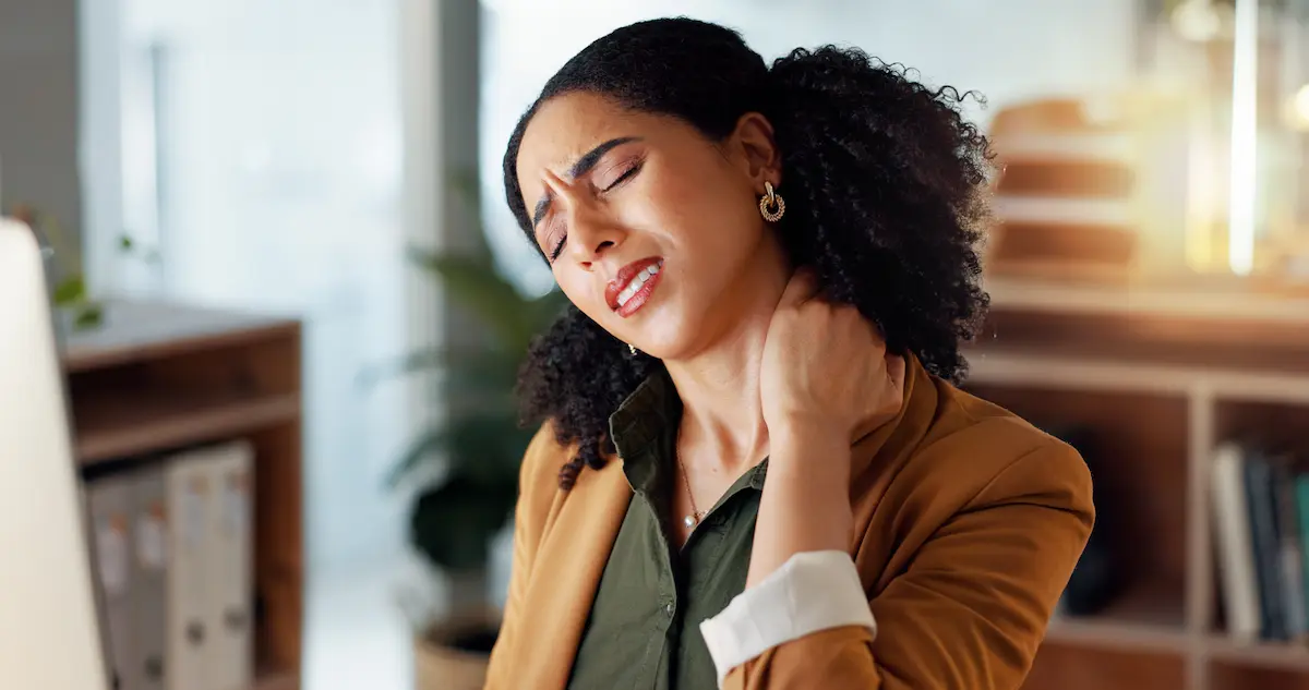A woman with curly black hair pulled back looks uncomfortable, closing her eyes and wincing while massaging the back of her neck with her hand. She is wearing a brown blazer over an olive green shirt in a brightly lit office setting.