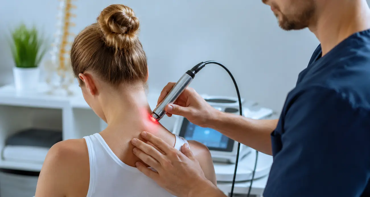 A practitioner applying cold laser therapy to the back of a patient's neck, with a visible red light glowing on the skin in a clinic setting.