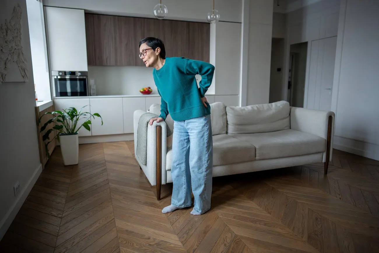 A woman standing in a modern living room, holding her lower back with one hand while leaning against the arm of a white sofa, appearing to be in pain.