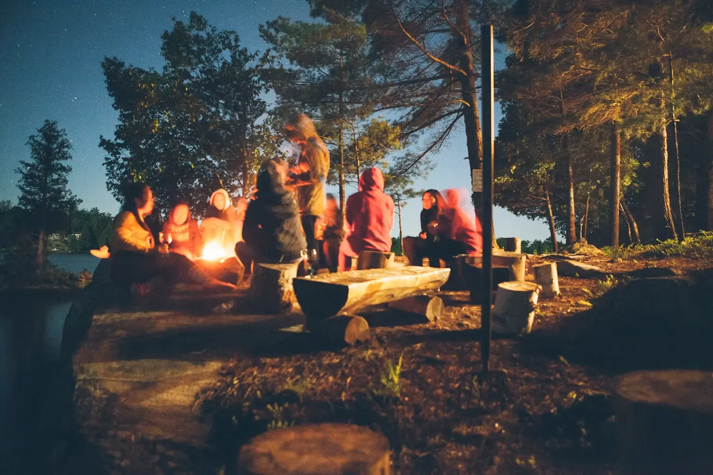 Group of people gathered around a campfire at night in a wooded area near a lake.