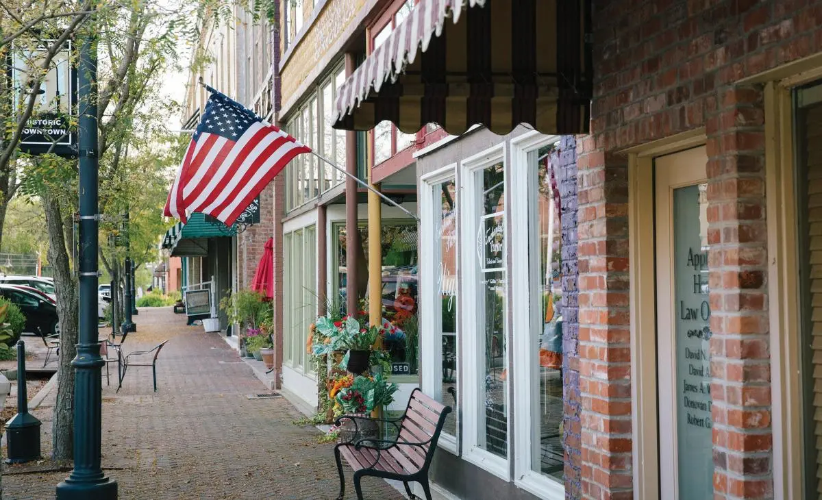 Historic downtown main street sidewalk lined with brick buildings, storefront windows, striped awnings, and an American flag hanging over the walkway.