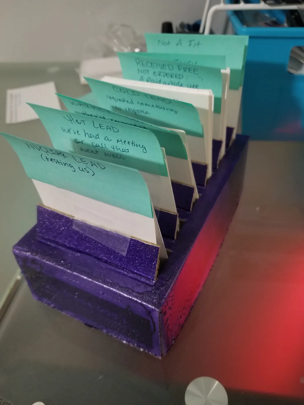 A cardboard box painted purple with glitter and patched with tape, holding white index cards behind hand-lettered mint-green dividers. The visible labels read: Nuclear Lead (testing us); Hot Lead — we've had a meeting or call that went well; Warm; Cold Lead — requested something, no response; Received free, not ordered; Not A Fit.