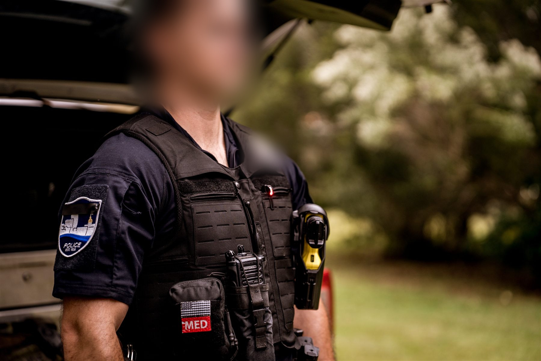 Patrol officer in a black Armor Express duty vest with radio, med pouch and body-worn gear, standing beside his open police vehicle
