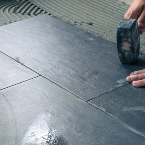 Une personne utilise un marteau en caoutchouc pour tapoter un carreau de sol gris foncé afin de le mettre en place sur un mortier frais.