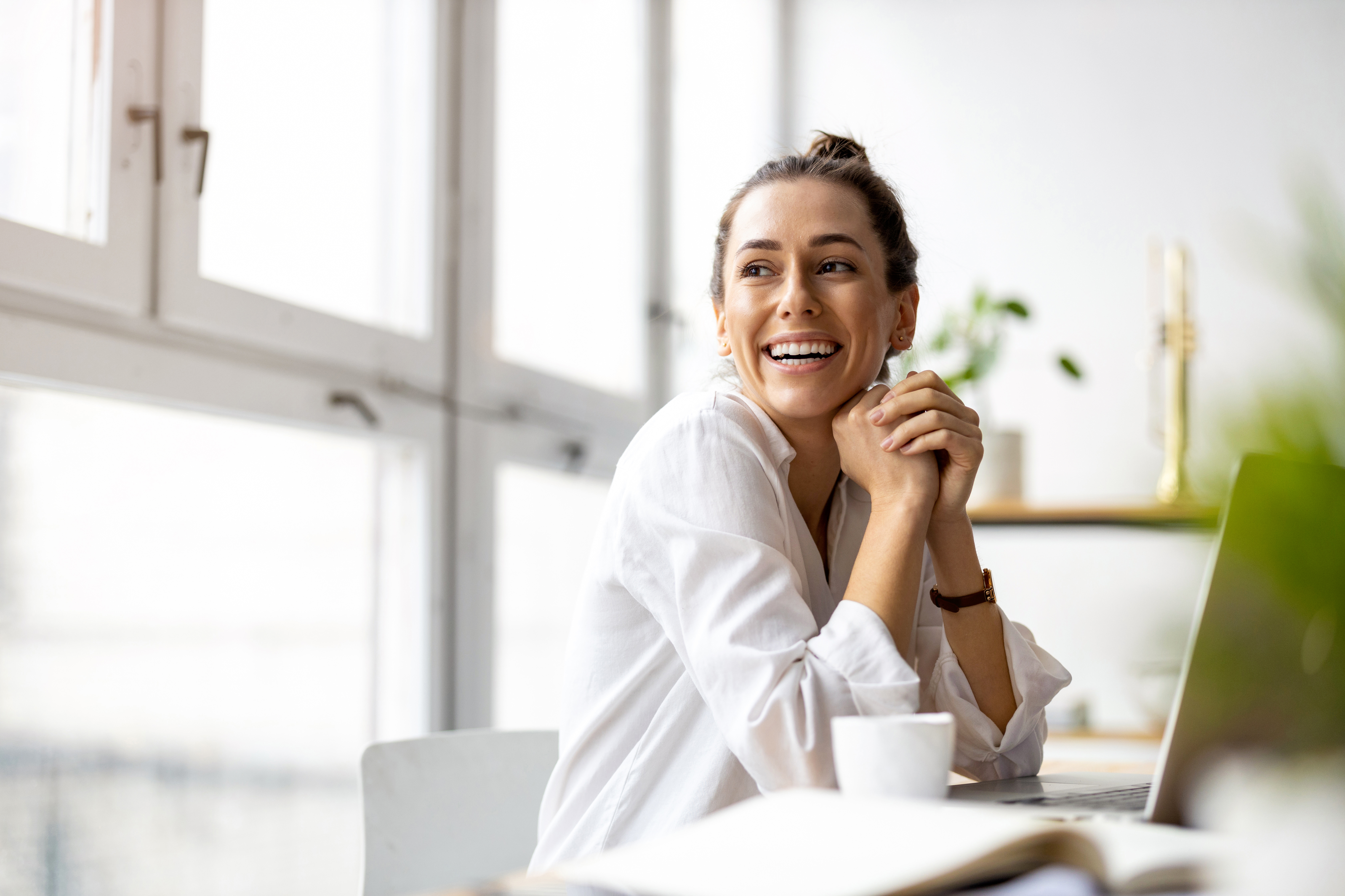 Smiling woman with hands clasped near her face, seated at a table with windows behind her, representing thoughtful integration