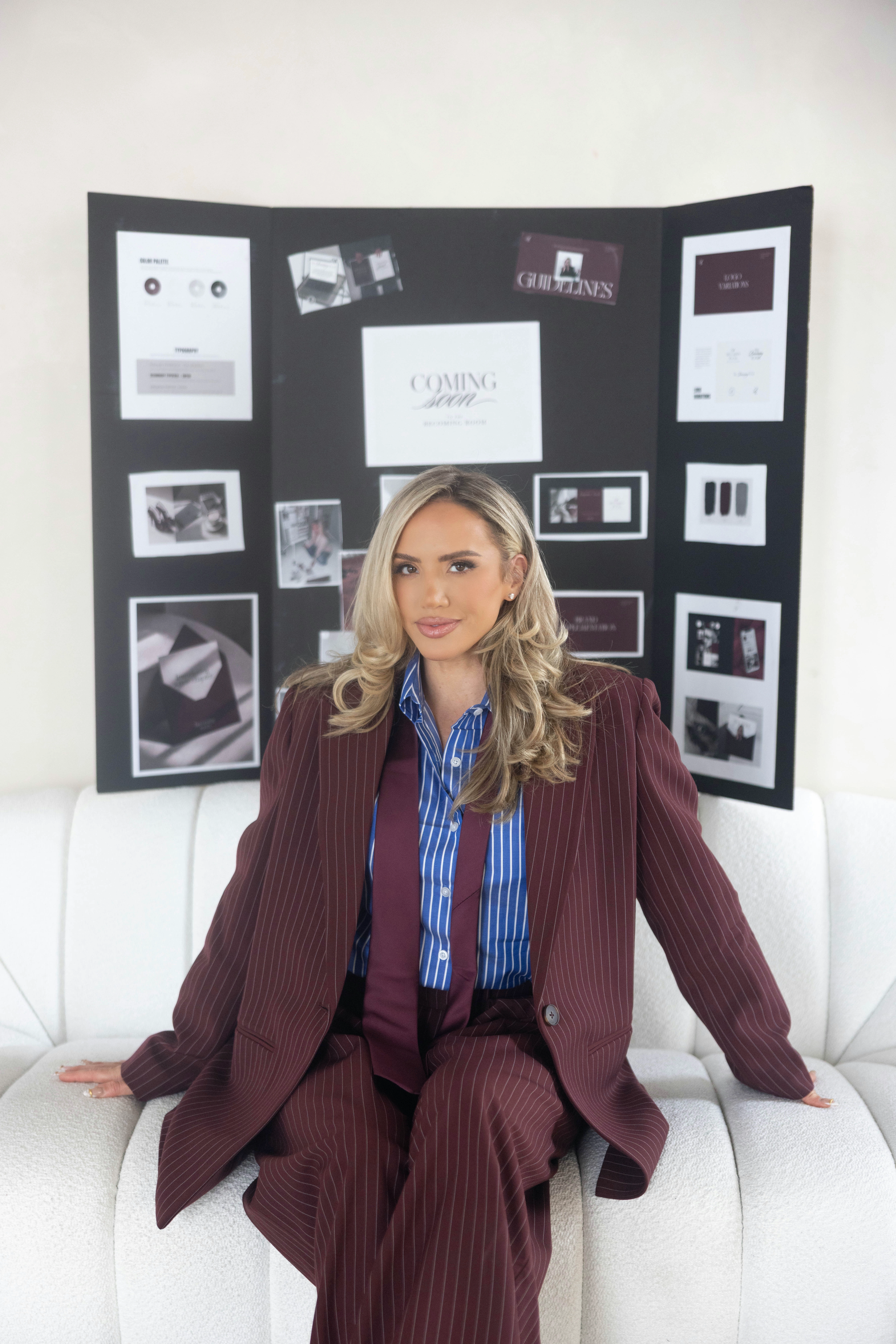Calli Wolff in a maroon pinstripe suit and blue striped shirt, seated on a white sofa in front of her brand vision board