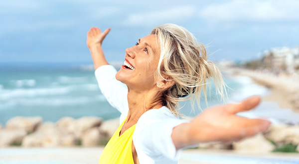 A happy middle-aged woman by the coast, looking up with arms wide open