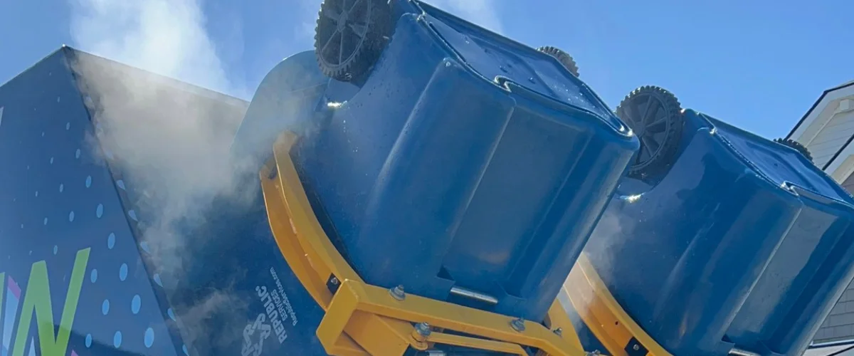 Two blue recycling bins being emptied into a collection truck, with steam or mist coming from the connection. Two blue recycling bins being emptied into a collection truck, with steam or mist coming from the connection.