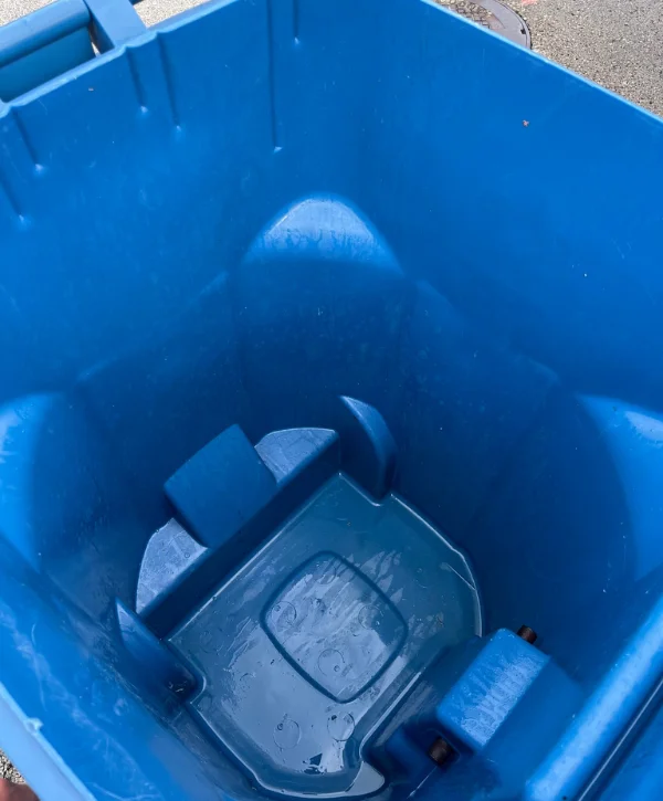 An overhead view looking down into an empty, wet blue plastic recycling bin with internal structural features.