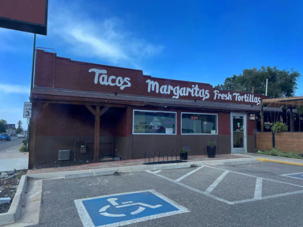 A small, brown brick restaurant with a sign reading "Tacos, Margaritas, Fresh Tortillas" above the entrance. The building features a large window next to the entrance door, with an "OPEN" sign visible inside. The exterior includes a ramp leading to the entrance, and there is a handicapped parking space in front of the building. The sky above is clear and blue.