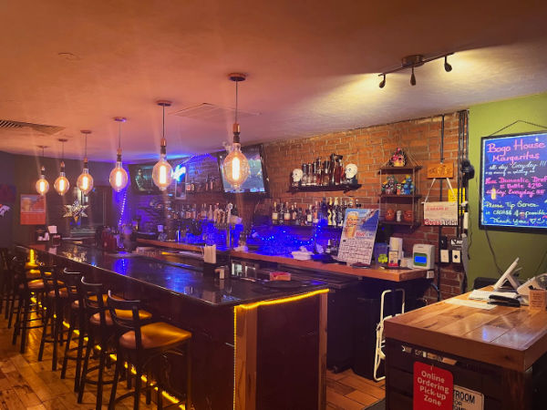 The interior of a cozy bar area with a long counter, lined with high-back chairs. The bar is well-lit with hanging pendant lights, creating a warm and inviting atmosphere. Behind the bar, shelves display various bottles of liquor, glassware, and a chalkboard menu advertising "Bogo House Margaritas." The walls feature a combination of exposed brick and painted surfaces, adding to the rustic charm.