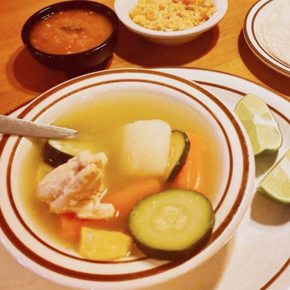 A bowl of caldo de pollo, a traditional Mexican chicken soup, with a clear broth containing chunks of tender chicken, zucchini, carrots, and a piece of potato. The bowl is served with a side of rice, a small bowl of salsa, and several lime wedges on a plate. In the background, there are warm tortillas ready to be served.