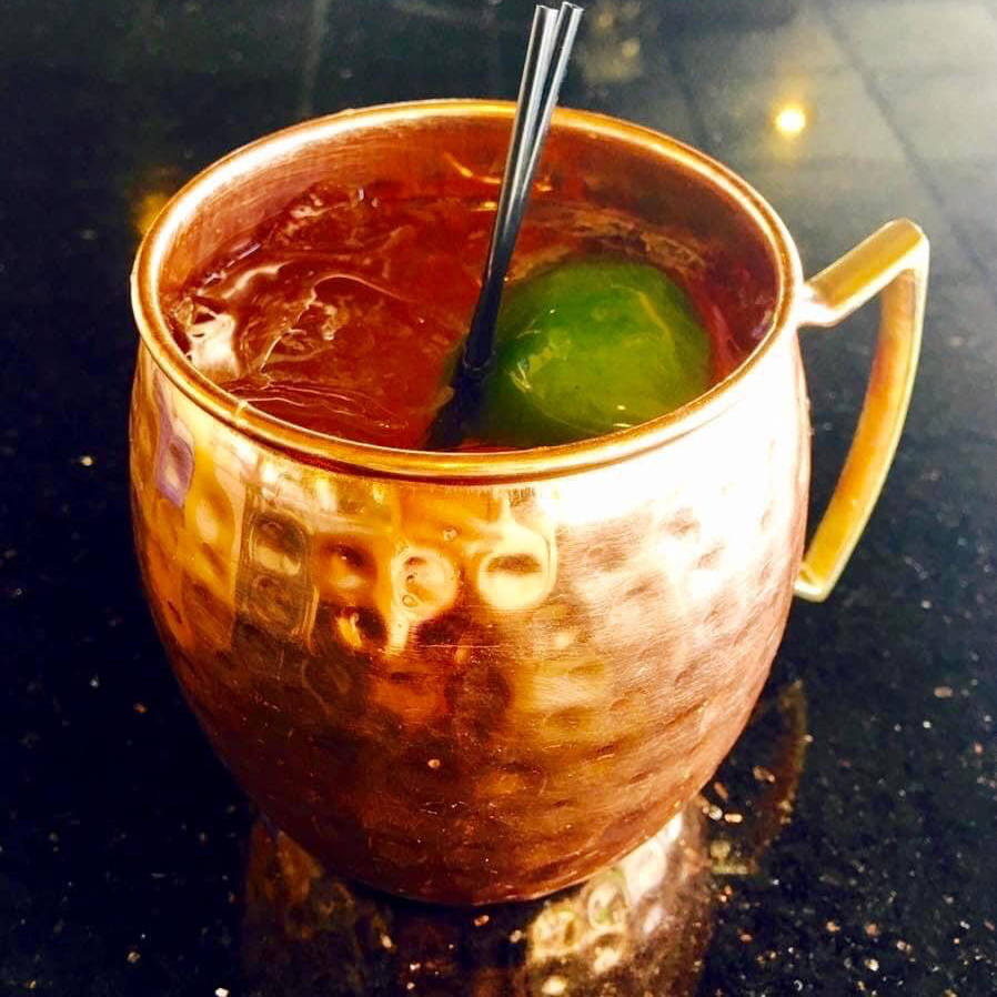 A Moscow Mule cocktail served in a hammered copper mug, garnished with a lime wedge. The drink is chilled with ice and has a refreshing appearance against a dark, glossy surface.