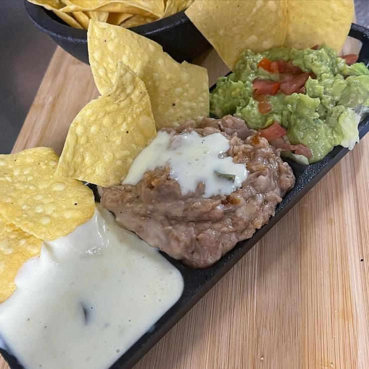 A tray with three compartments featuring a serving of tortilla chips paired with queso dip, refried beans topped with melted cheese, and freshly made guacamole mixed with diced tomatoes and onions.