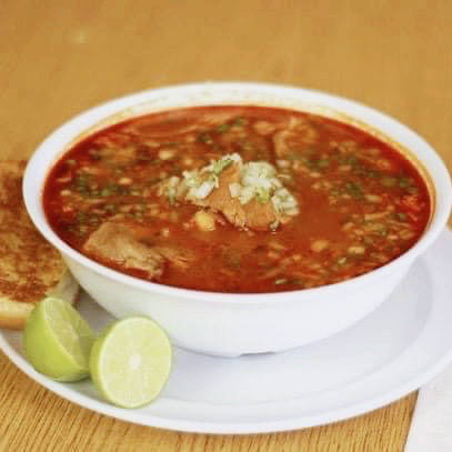 A bowl of traditional menudo, a Mexican soup made with a red, savory broth and tripe, garnished with chopped onions and cilantro. The bowl is served on a white plate alongside two lime wedges and a toasted slice of bolillo bread, placed on a wooden table.