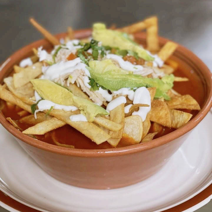 A bowl of tortilla soup topped with crispy tortilla strips, shredded chicken, sliced avocado, and a drizzle of sour cream. The soup is served in a clay bowl, adding a rustic touch to the presentation.