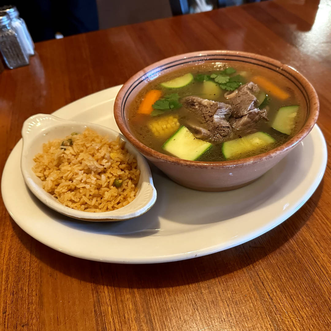 A bowl of caldo de res, a traditional Mexican beef soup, served with chunks of tender beef, zucchini, corn, and carrots in a clear broth, garnished with fresh cilantro. The bowl is accompanied by a side of Mexican-style rice, served in a small dish on the same plate.