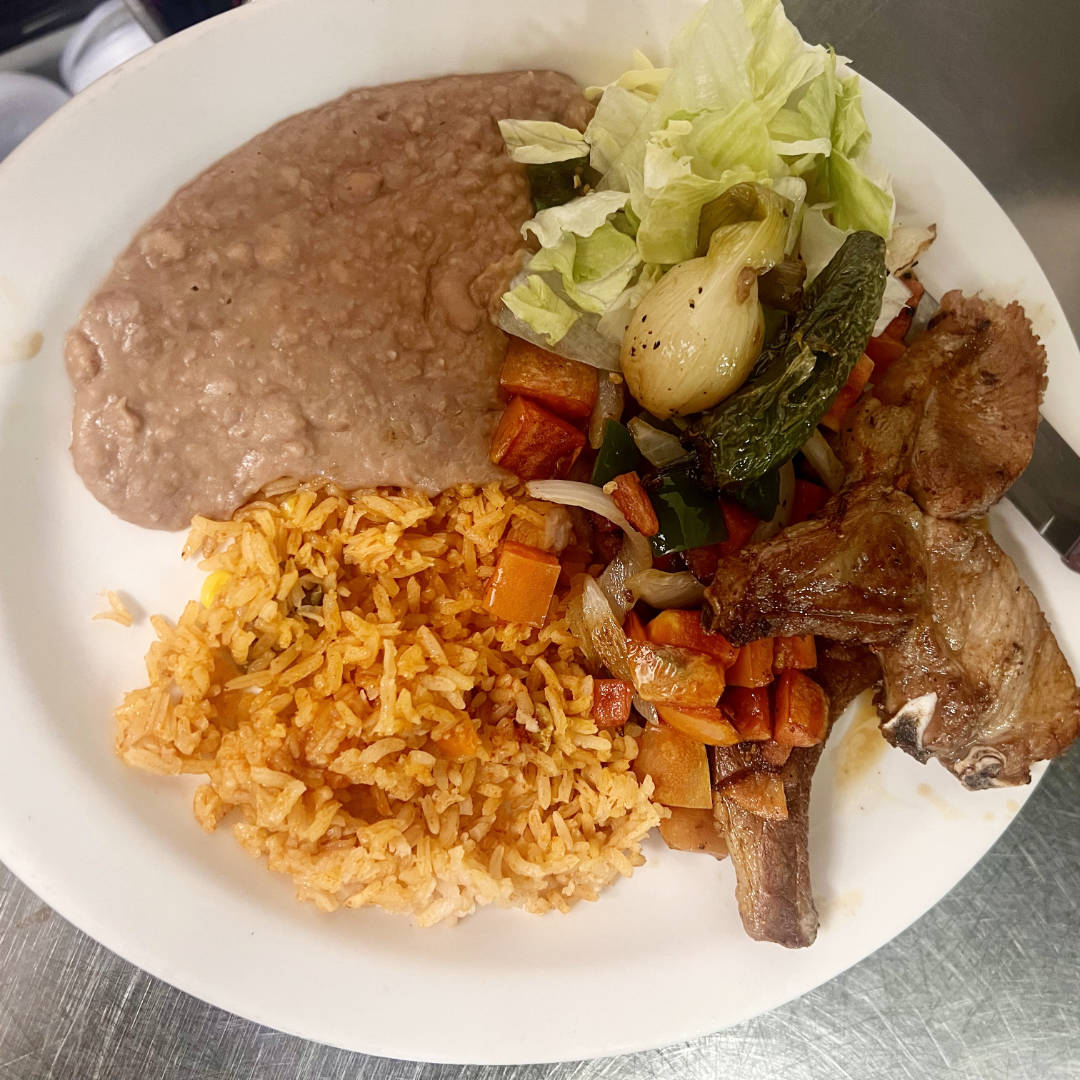 A plate of food featuring a serving of refried beans, Mexican-style rice, and grilled pork chops. The plate is also garnished with sautéed vegetables, including carrots, onions, and green peppers, alongside fresh lettuce and a roasted green onion.