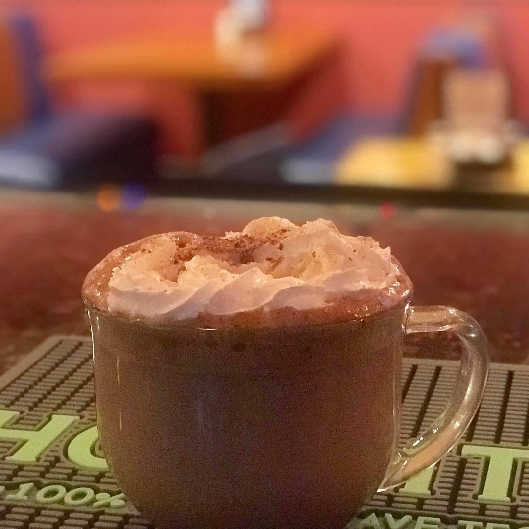 A warm beverage called Chocolate Mexicano Abuelita. It's a hot chocolate or a specialty coffee, served in a clear glass mug. The drink is topped with a generous amount of whipped cream and a sprinkle of cinnamon, giving it a cozy, indulgent appearance. The background is softly blurred, showing a warm, inviting atmosphere with muted tones.